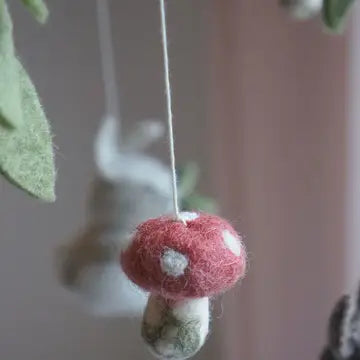 A close-up of a felted wool mushroom ornament hanging from a thin string, with a soft pink background and blurred decorative elements.
