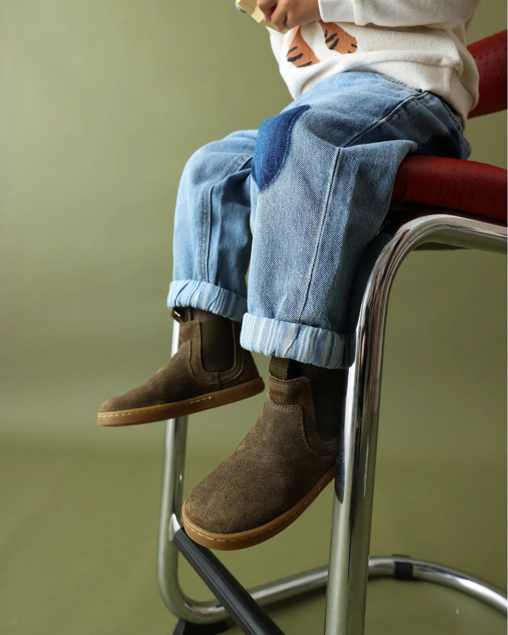 A child sitting on a stool wearing light blue jeans, brown suede boots, and a white top, photographed against a muted green background.