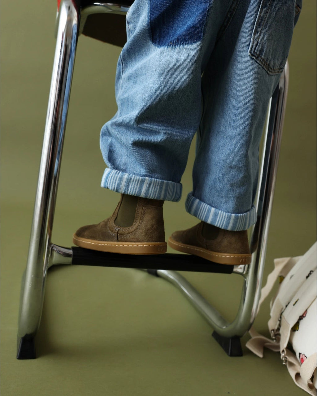 A person wearing blue jeans and brown boots stands on a metal step stool against a solid olive green background.