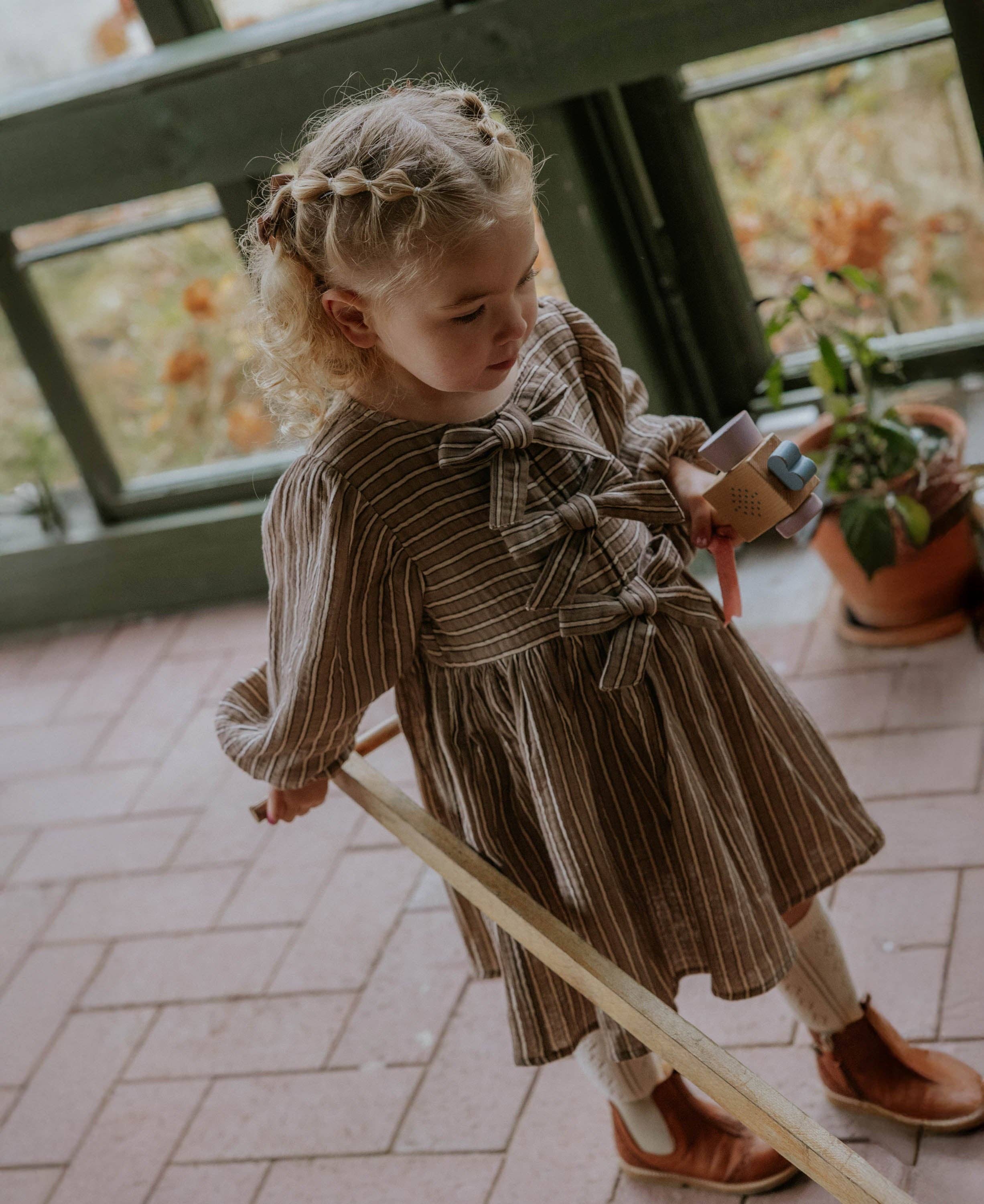 A young girl with blonde braided hair wearing a brown and beige vertically striped long-sleeve dress with a large bow at the waist, standing on a tiled porch next to a wooden railing.