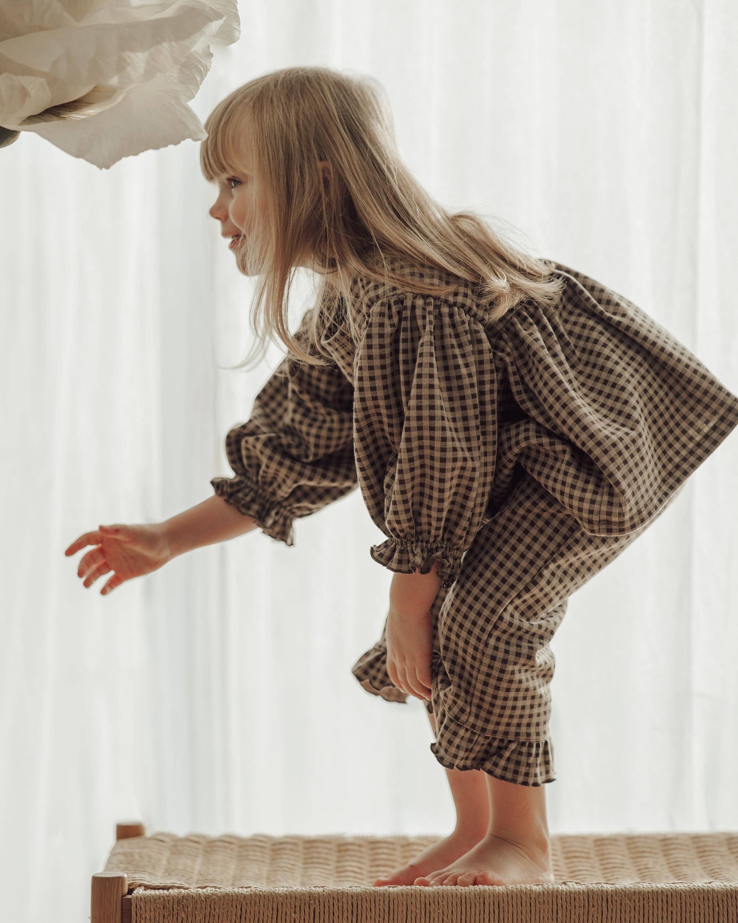 Child wearing a checkered outfit standing on a woven bench with a white curtain background