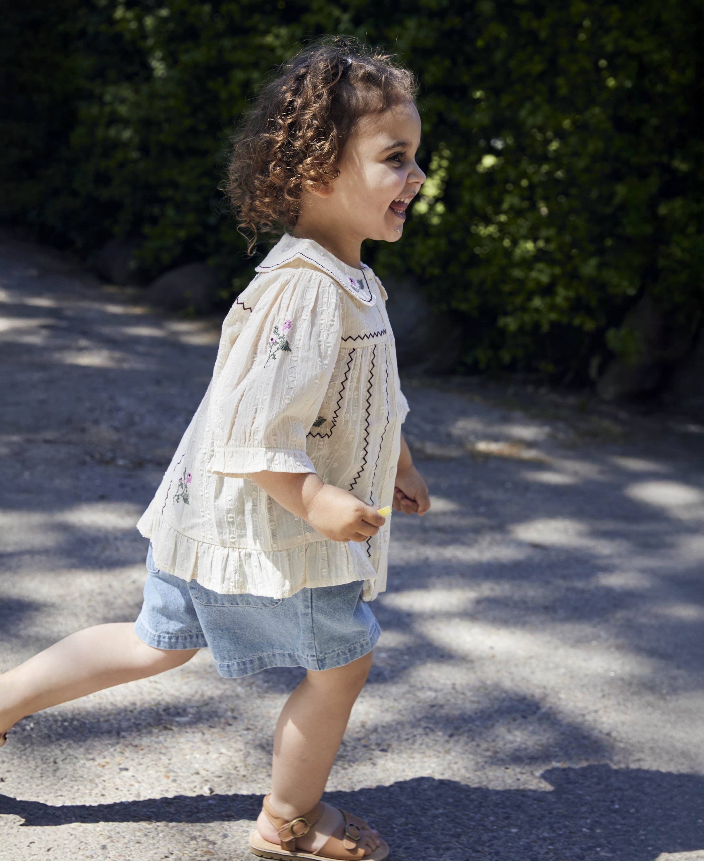 A toddler girl walking outdoors on a sunlit path, wearing a cream embroidered blouse with ruffled sleeves and denim shorts.