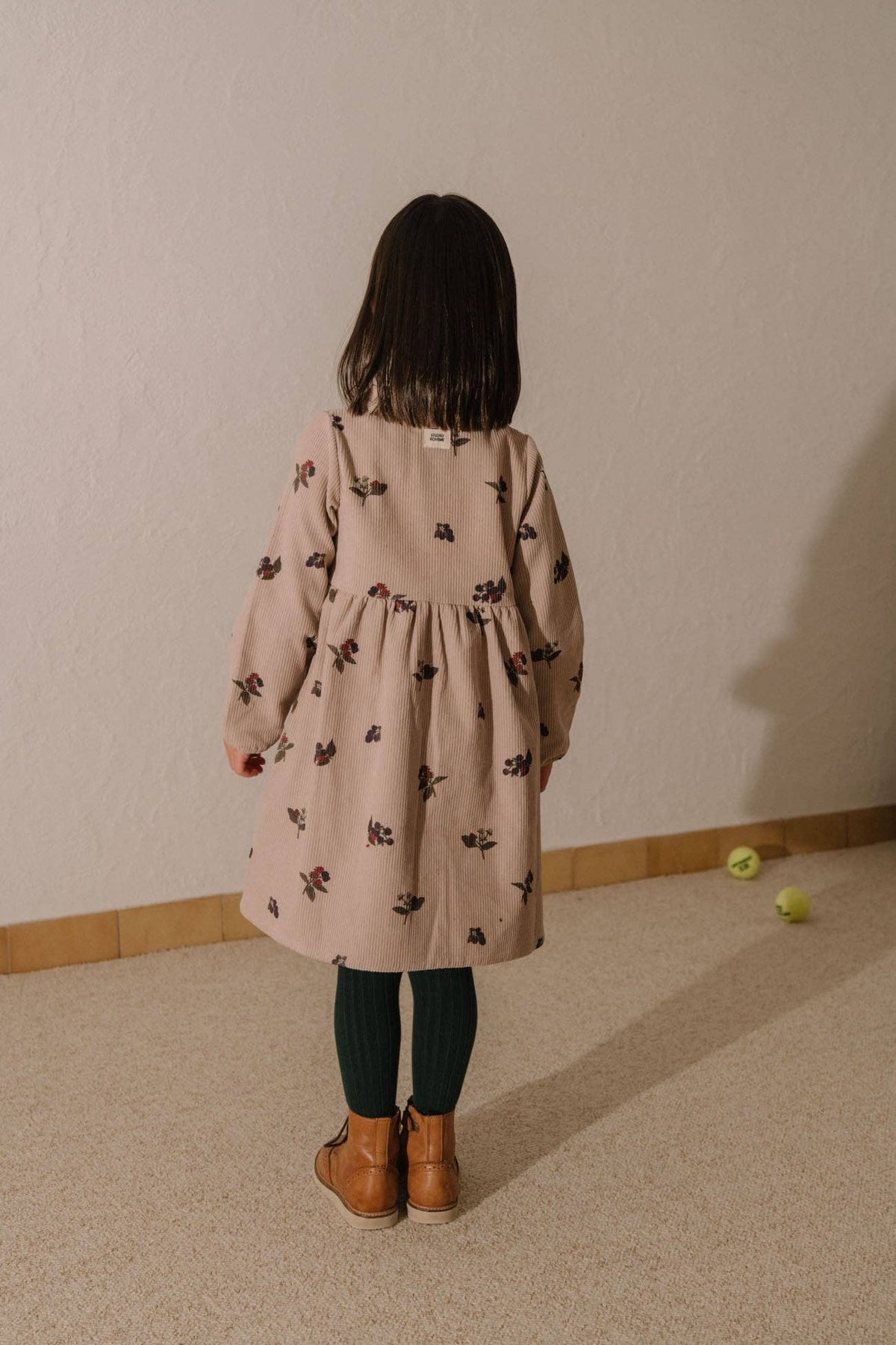 Child wearing a pink dress with a pattern, standing in a room with a plain wall and light-colored floor.