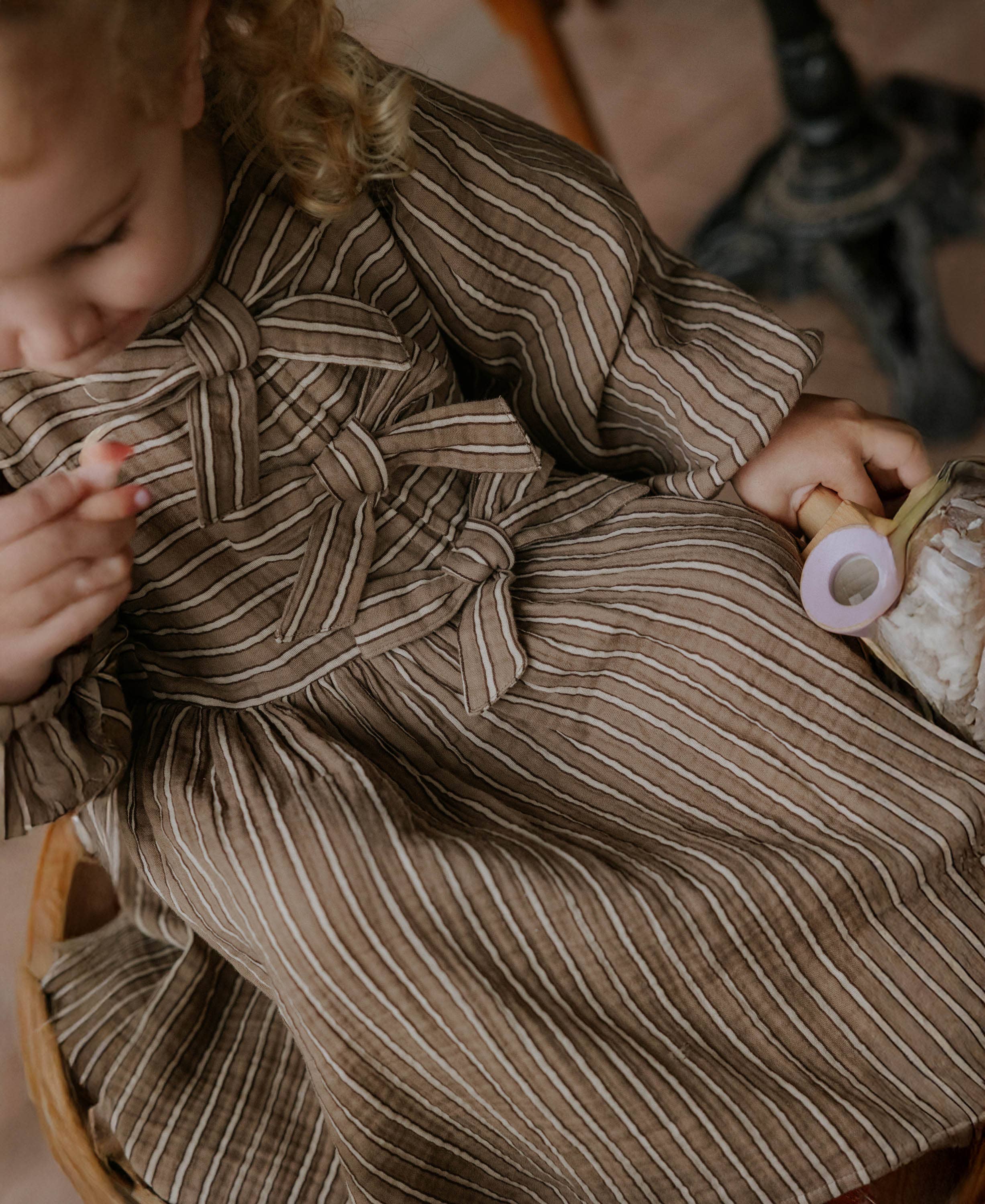 A close-up lifestyle shot of a toddler wearing a brown and beige vertically striped dress with a large front bow, sitting in a wooden chair and holding a pacifier clip.