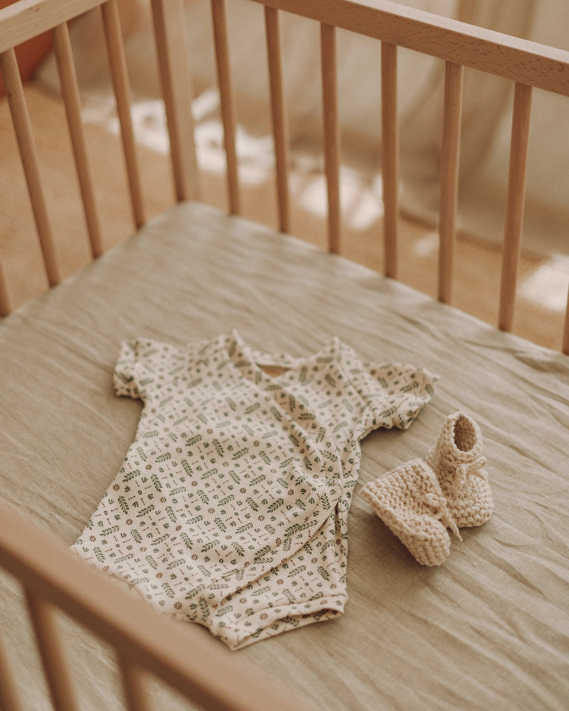 A baby outfit set consisting of a short-sleeve cream bodysuit with a green leaf pattern and matching cream knit booties, laid out on a beige mattress inside a wooden crib.
