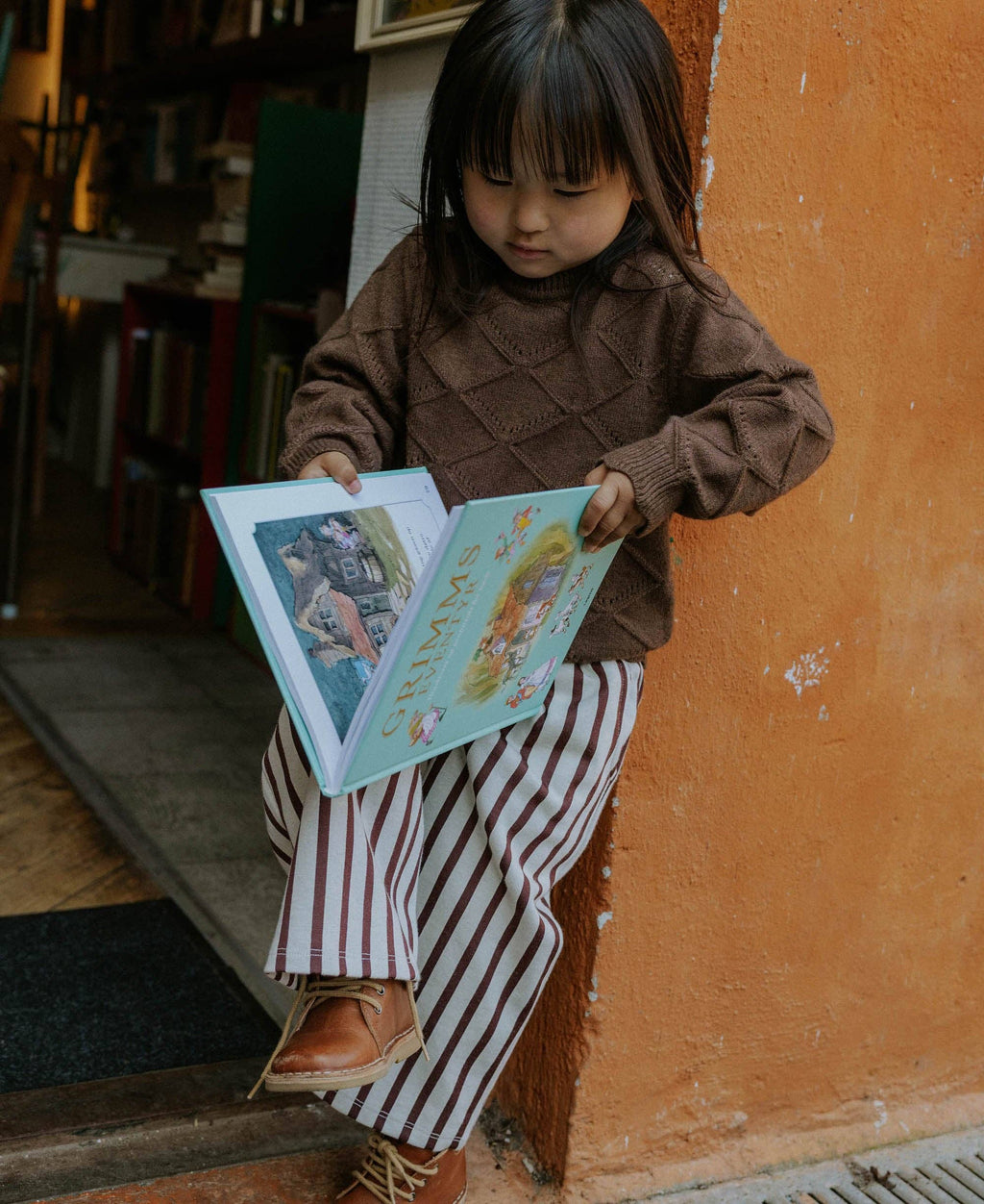 A young child sitting on a wooden step against an orange wall, reading a colorful book. The child is wearing a brown knit sweater and red-and-white vertically striped pants.