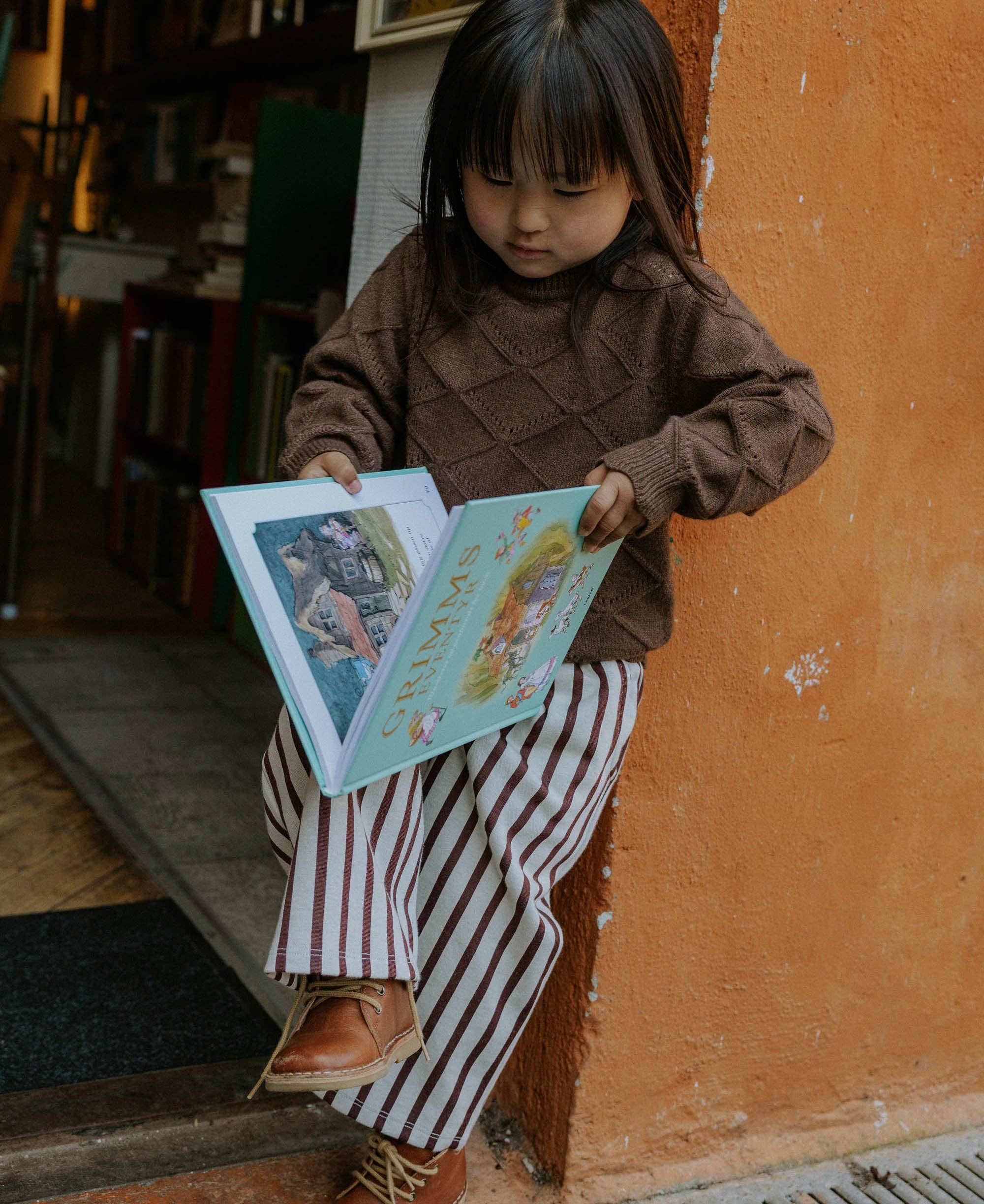 A young child sitting on a wooden step against an orange wall, reading a colorful book. The child is wearing a brown knit sweater and red-and-white vertically striped pants.