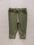 Olive green knit baby leggings with a matching drawstring waist, laid flat on a textured beige linen background.