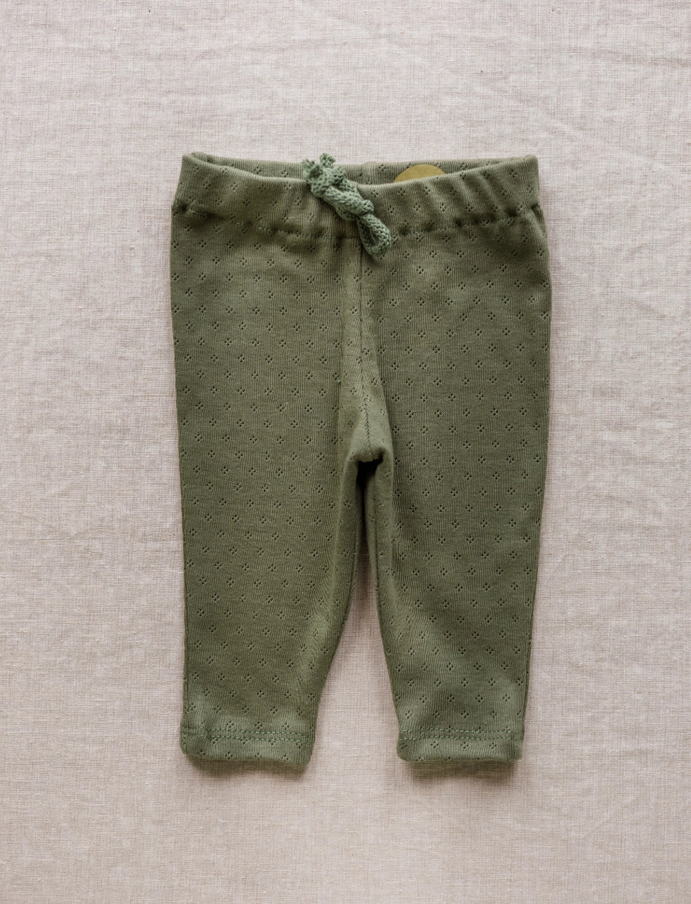 Olive green knit baby leggings with a matching drawstring waist, laid flat on a textured beige linen background.