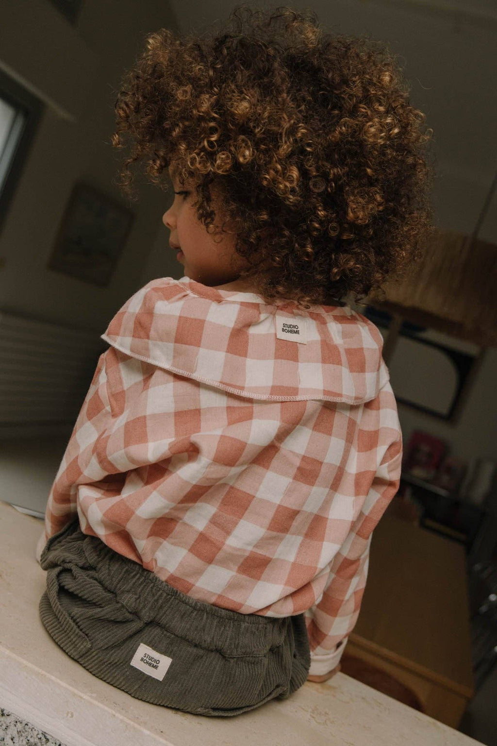 Child wearing a checkered shirt and gray pants standing on a step indoors.