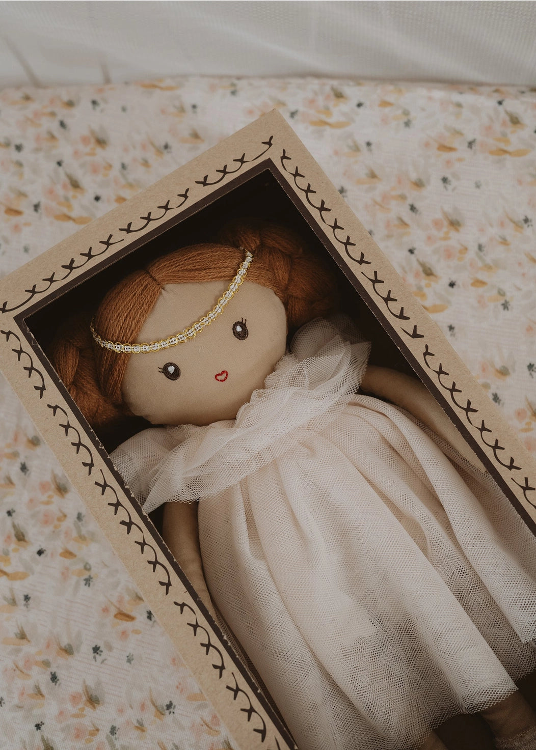 Small doll with a white dress and red hair in a decorative box with floral patterns.