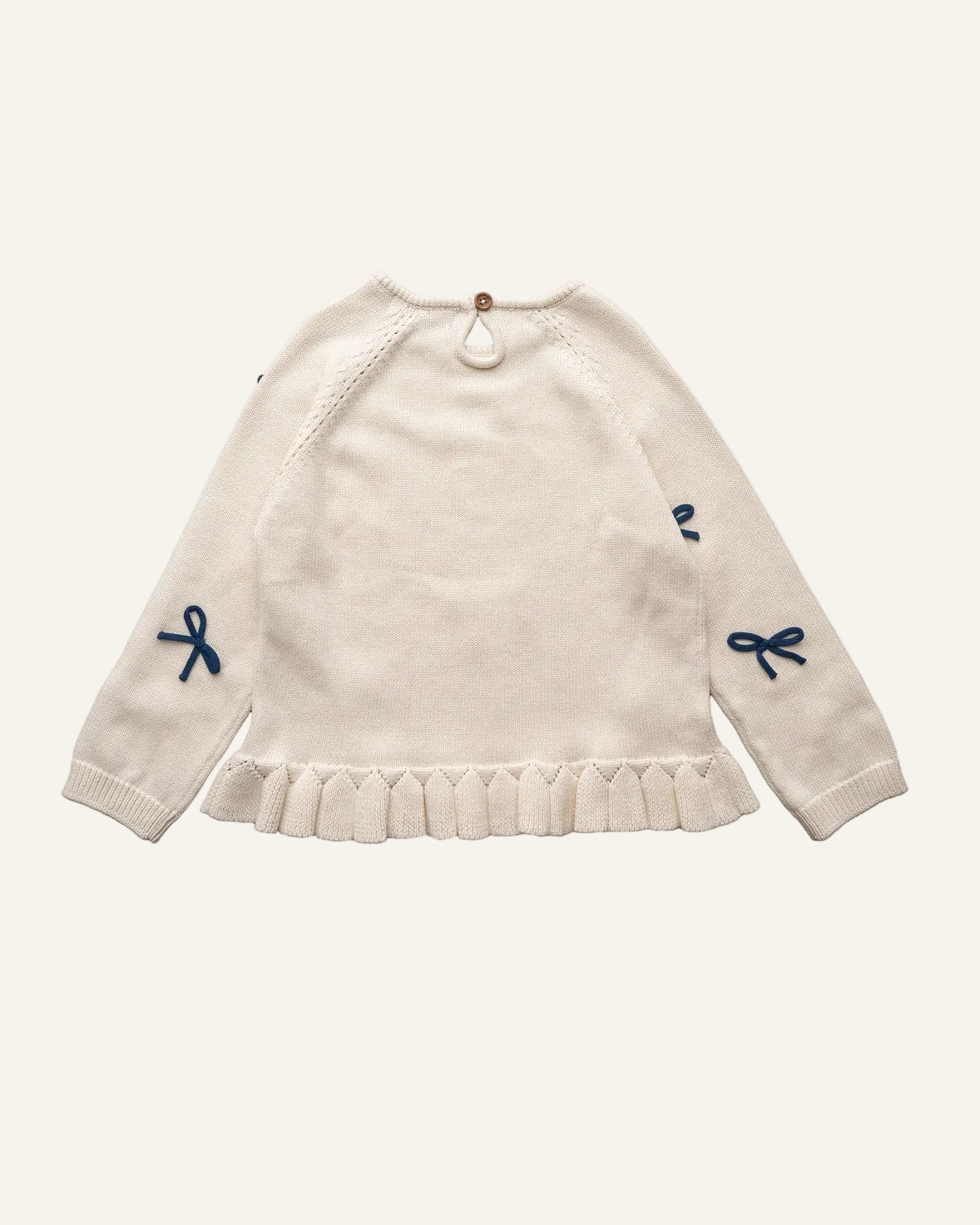 An off-white knit sweater for toddlers, featuring a ruffled hem, raglan sleeves, and small navy blue bow details on the sleeves, shown against a plain white background.