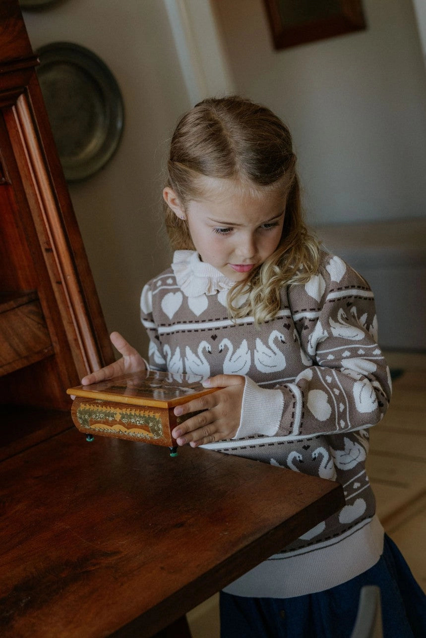 A young girl with blonde hair looking down at a small wooden box on a dark wooden table, wearing a brown and white patterned sweater with swan motifs.