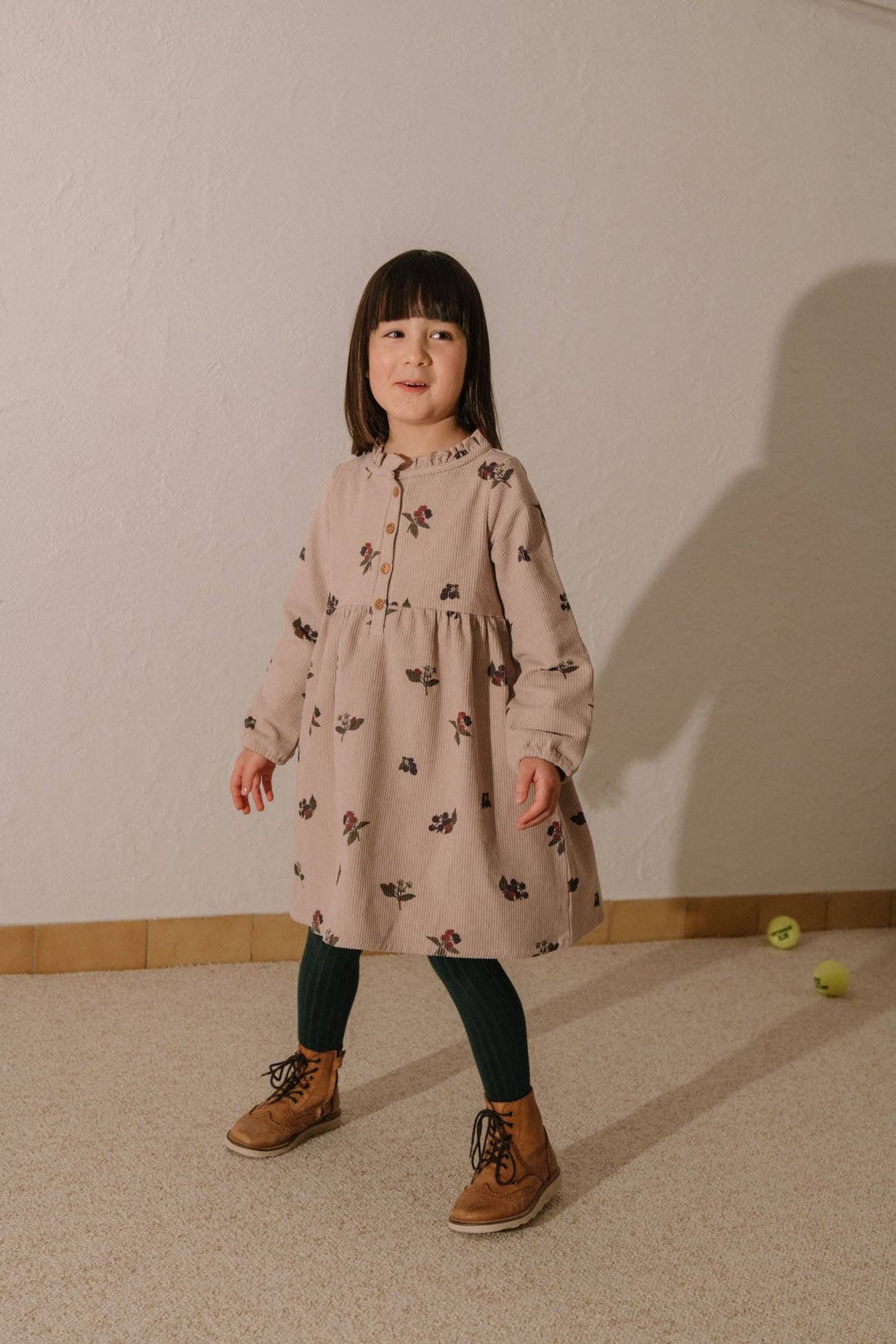 Young girl in a patterned dress standing against a plain wall with a shadow on the floor.