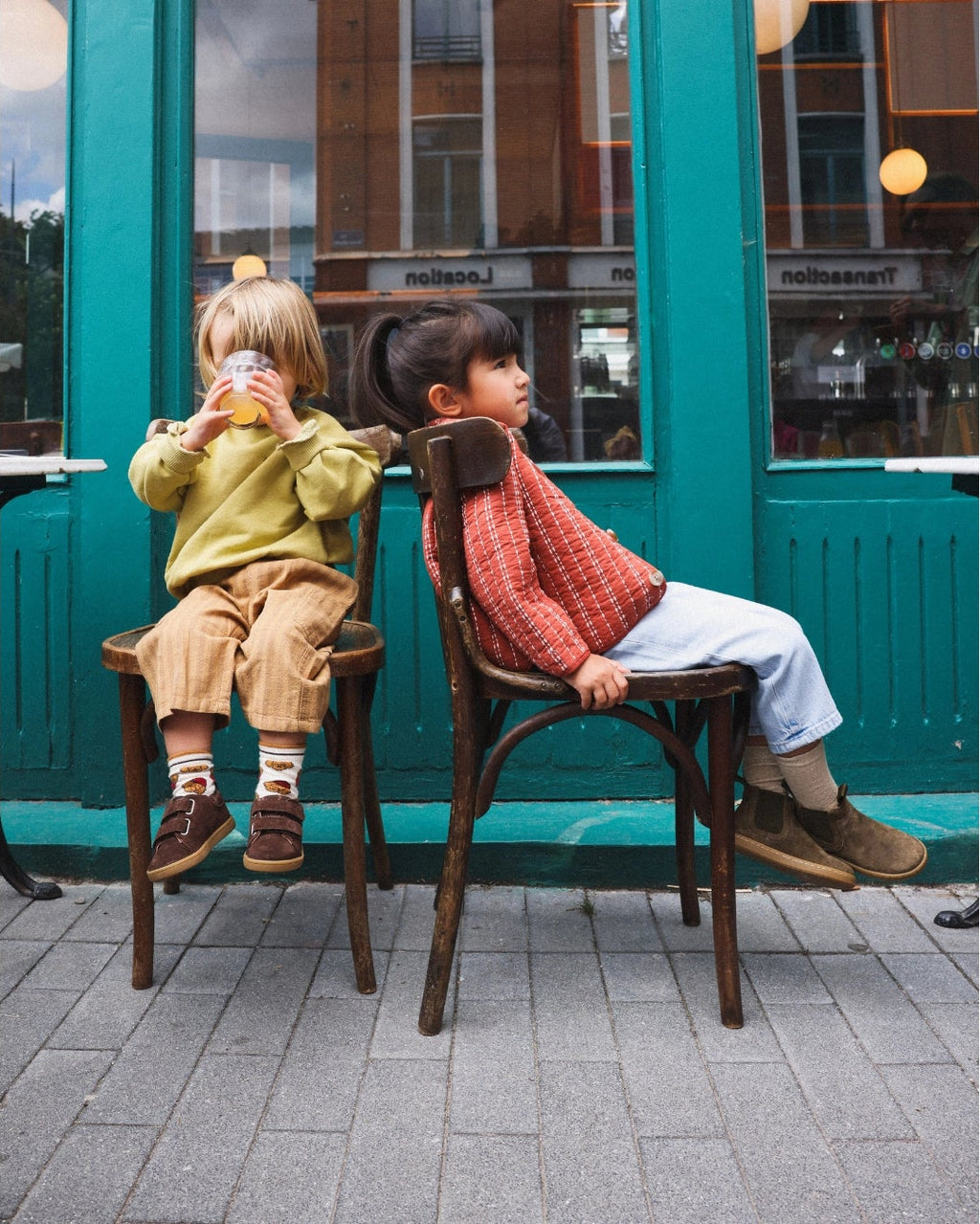 Two children sitting on dark wooden chairs outside a teal storefront, wearing cozy autumn clothing. The scene is set on a paved outdoor seating area with a teal building in the background.