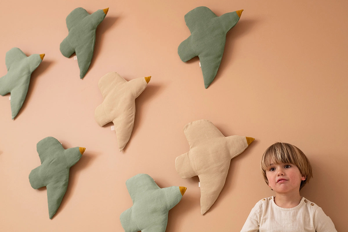 A child stands beside a peach-colored wall decorated with six bird-shaped plush pillows in sage green and cream, each with a small yellow beak.