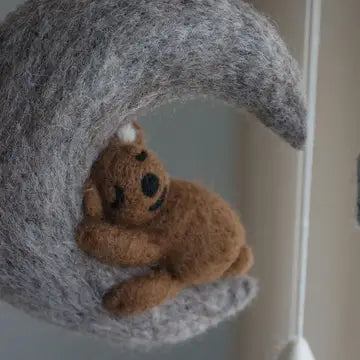 A small brown felt teddy bear with black embroidered eyes and mouth, nestled inside a crescent-shaped gray felt moon decoration.