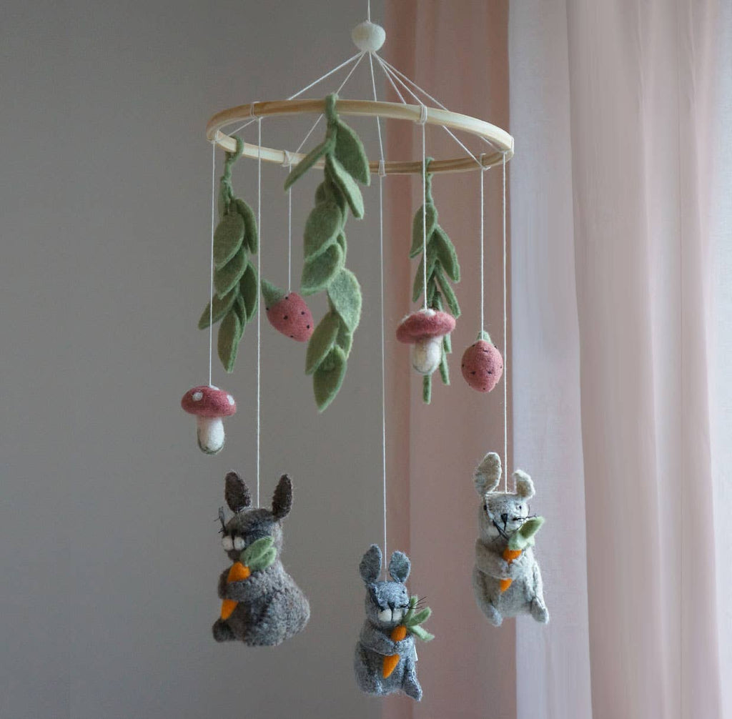 A felt baby mobile with three gray rabbits holding carrots, green leaves, pink and white mushrooms, and a light wood hanging ring, shown against a soft pink curtain and pale wall.