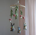 A felt baby mobile with three gray rabbits holding carrots, green leaves, pink and white mushrooms, and a light wood hanging ring, shown against a soft pink curtain and pale wall.