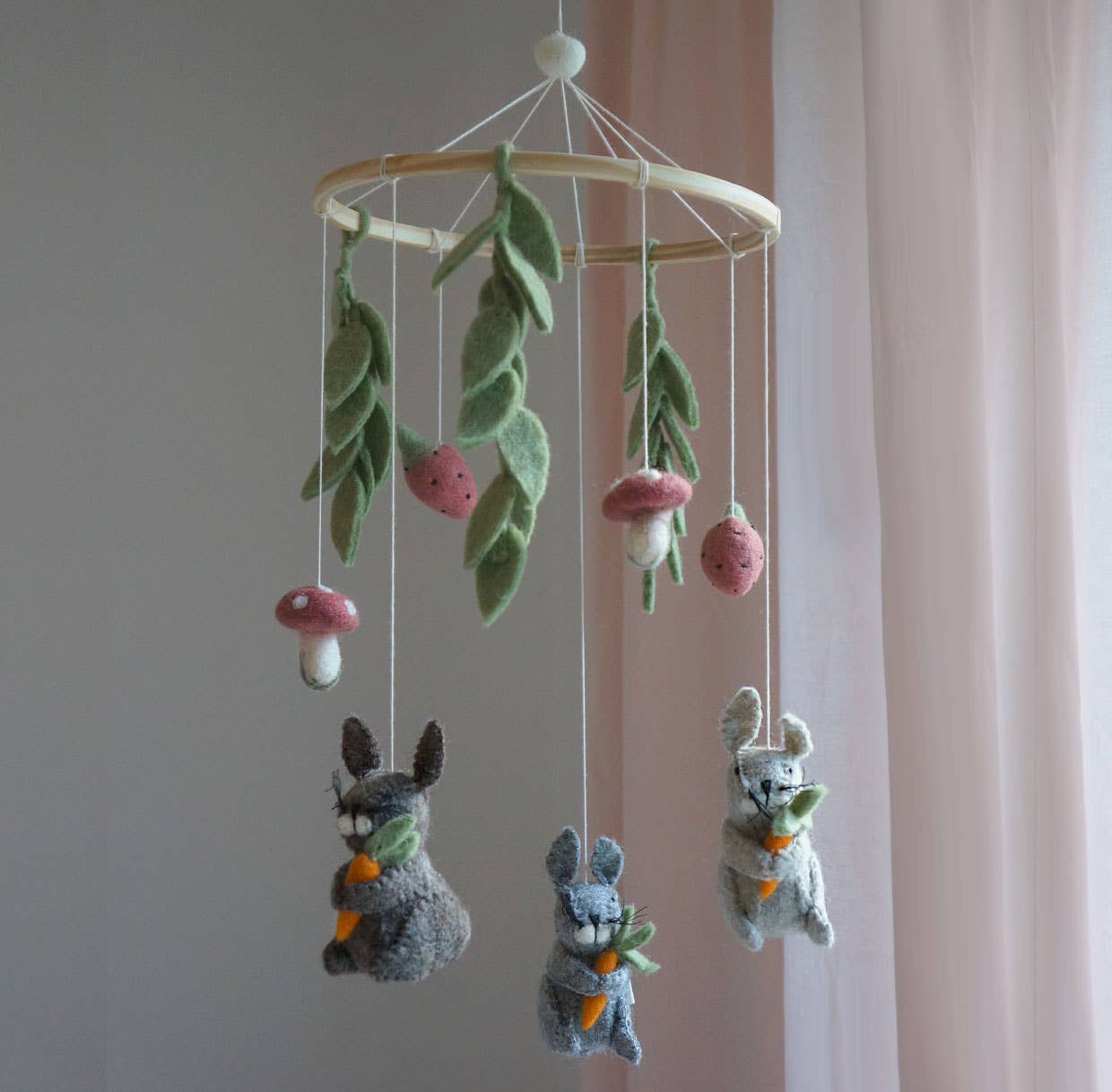 A felt baby mobile with three gray rabbits holding carrots, green leaves, pink and white mushrooms, and a light wood hanging ring, shown against a soft pink curtain and pale wall.