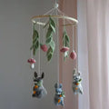 A felt baby mobile hanging from a wooden hoop, featuring green leaves, pink mushrooms, and three small rabbit figures in gray, white, and blue-gray holding carrots, set against a soft pink and gray background.
