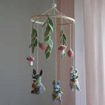A felt baby mobile hanging from a wooden hoop, featuring green leaves, pink mushrooms, and three small rabbit figures in gray, white, and blue-gray holding carrots, set against a soft pink and gray background.