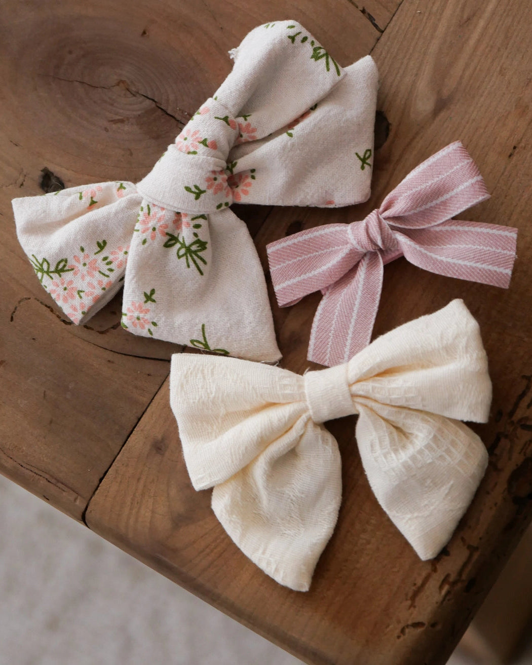 Three fabric hair bows arranged on a rustic wooden surface: one cream bow with a small pink floral print, one pale pink striped bow, and one cream textured bow.