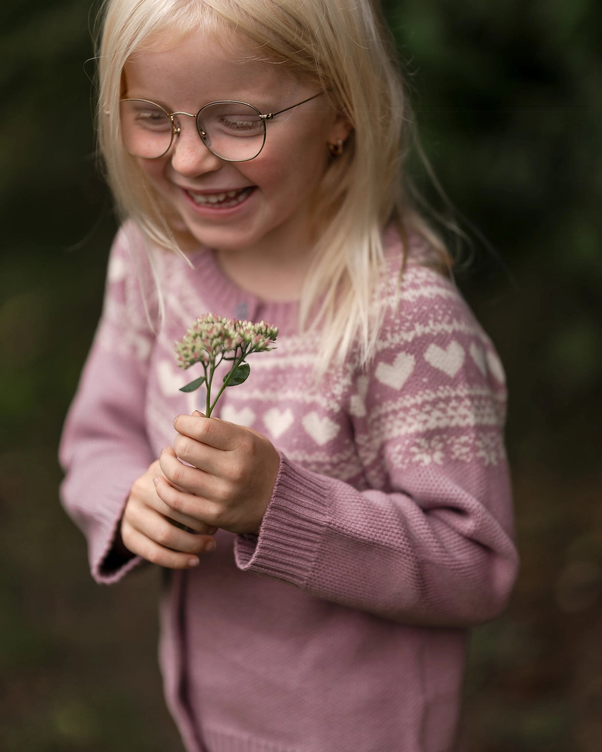 A young blonde girl with glasses smiling while holding a small sprig of wildflowers, wearing a dusty pink knit sweater with white heart patterns, set against a soft, blurred green forest background.
