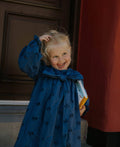 Child in a blue dress with a bow standing indoors against a red curtain.