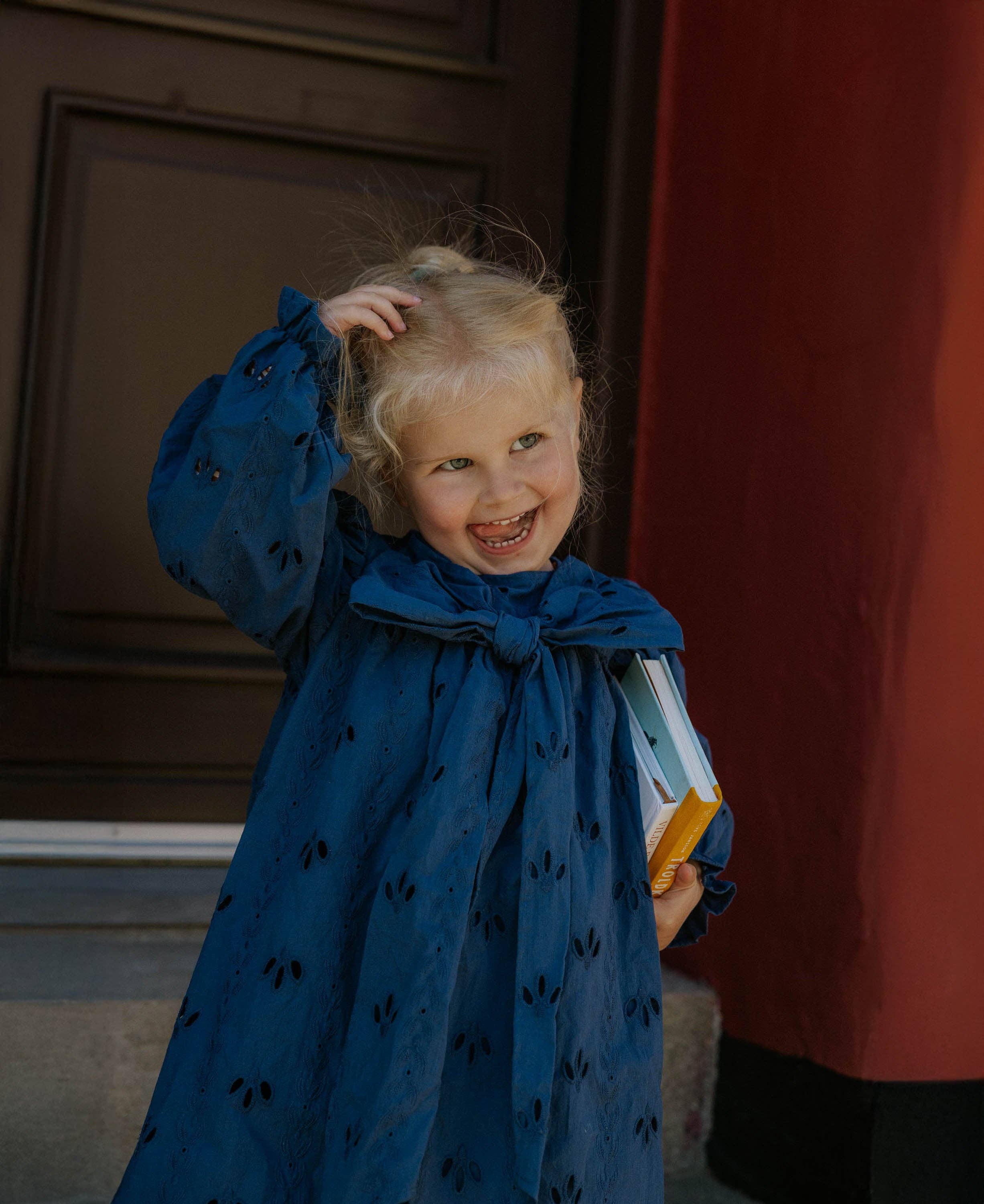 Child in a blue dress with a bow standing indoors against a red curtain.