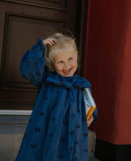 Child in a blue dress with a bow standing indoors against a red curtain.