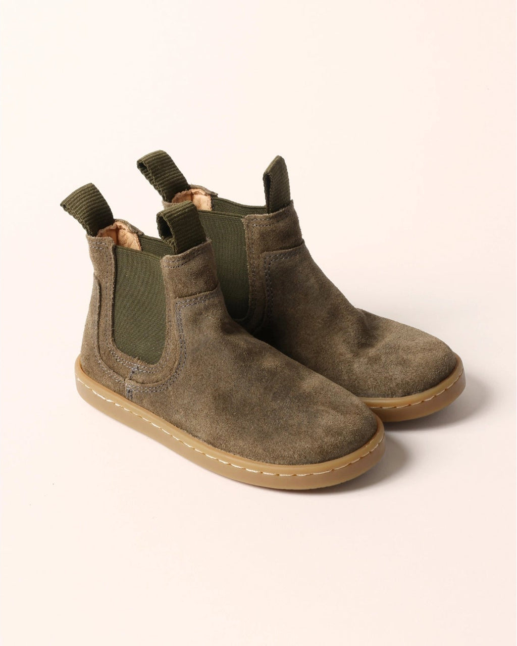 A pair of olive green suede Chelsea boots with elastic side panels and tan soles, displayed on a light beige background.