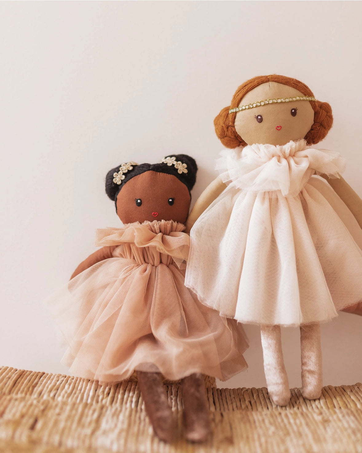 Two handmade fabric dolls dressed in elegant dresses, standing on a woven straw mat against a soft beige background. One doll has dark brown skin and black hair with white flowers, wearing a peach tulle dress.