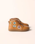 A pair of tan leather high-top boots decorated with colorful travel-style patches, shown against a solid pale peach background.