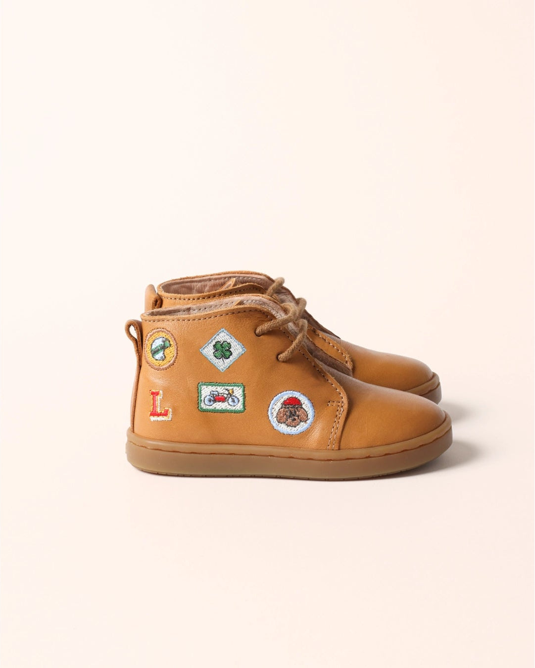 A pair of tan leather high-top boots decorated with colorful travel-style patches, shown against a solid pale peach background.