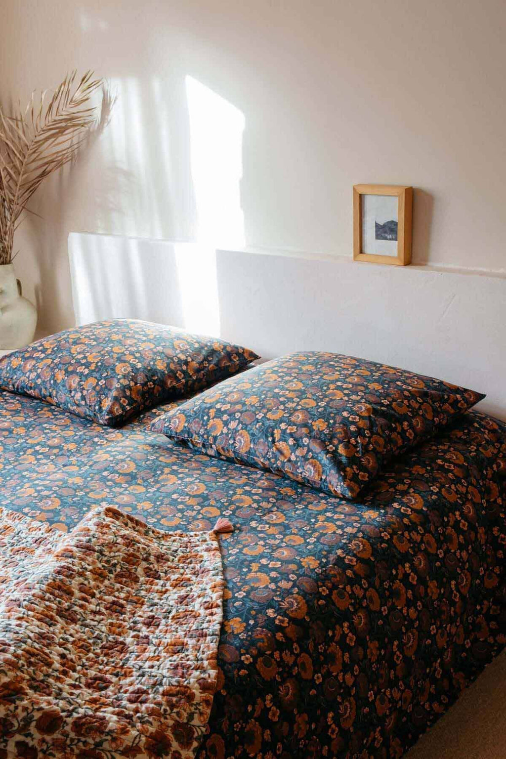 Bed with floral bedding in a room with a window and framed picture on the wall.