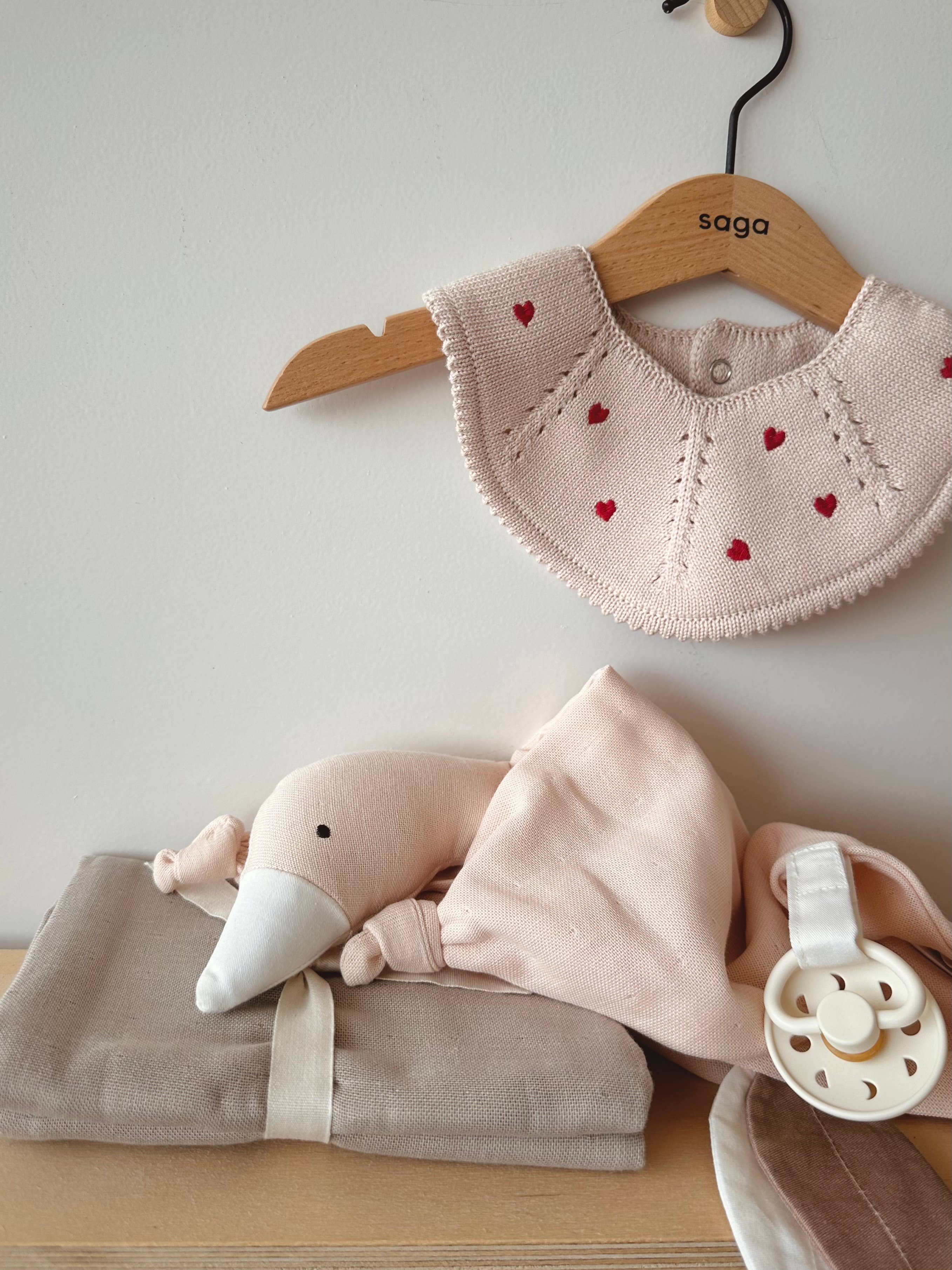 Baby bib with heart patterns, plush toy, and pacifier on a wooden surface with 'saga' branding.