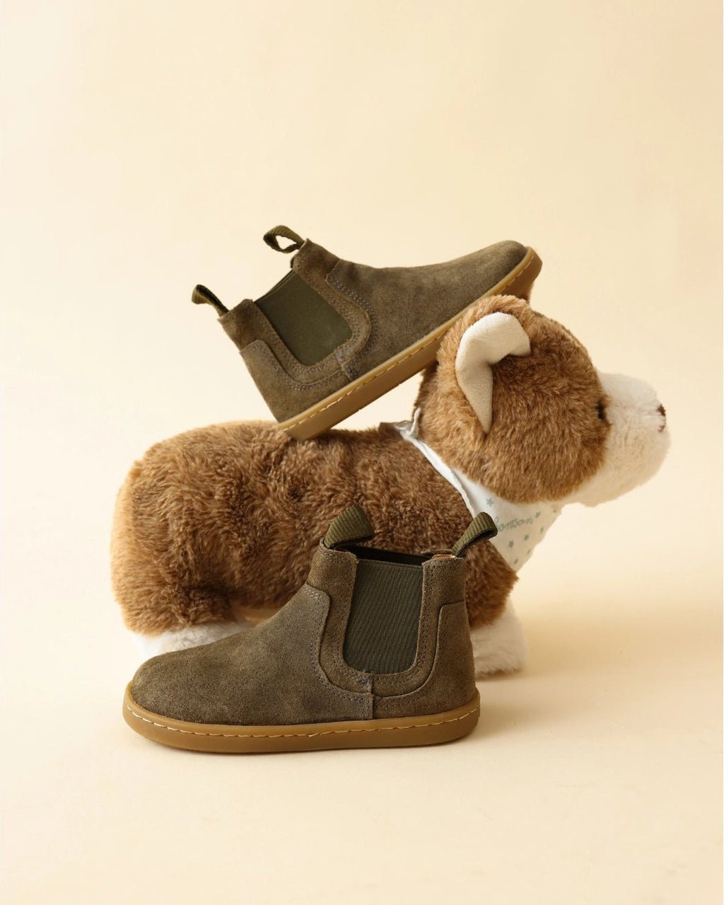 A pair of taupe-brown suede Chelsea-style ankle boots displayed on a plush brown and white dog toy, set against a solid warm beige background.