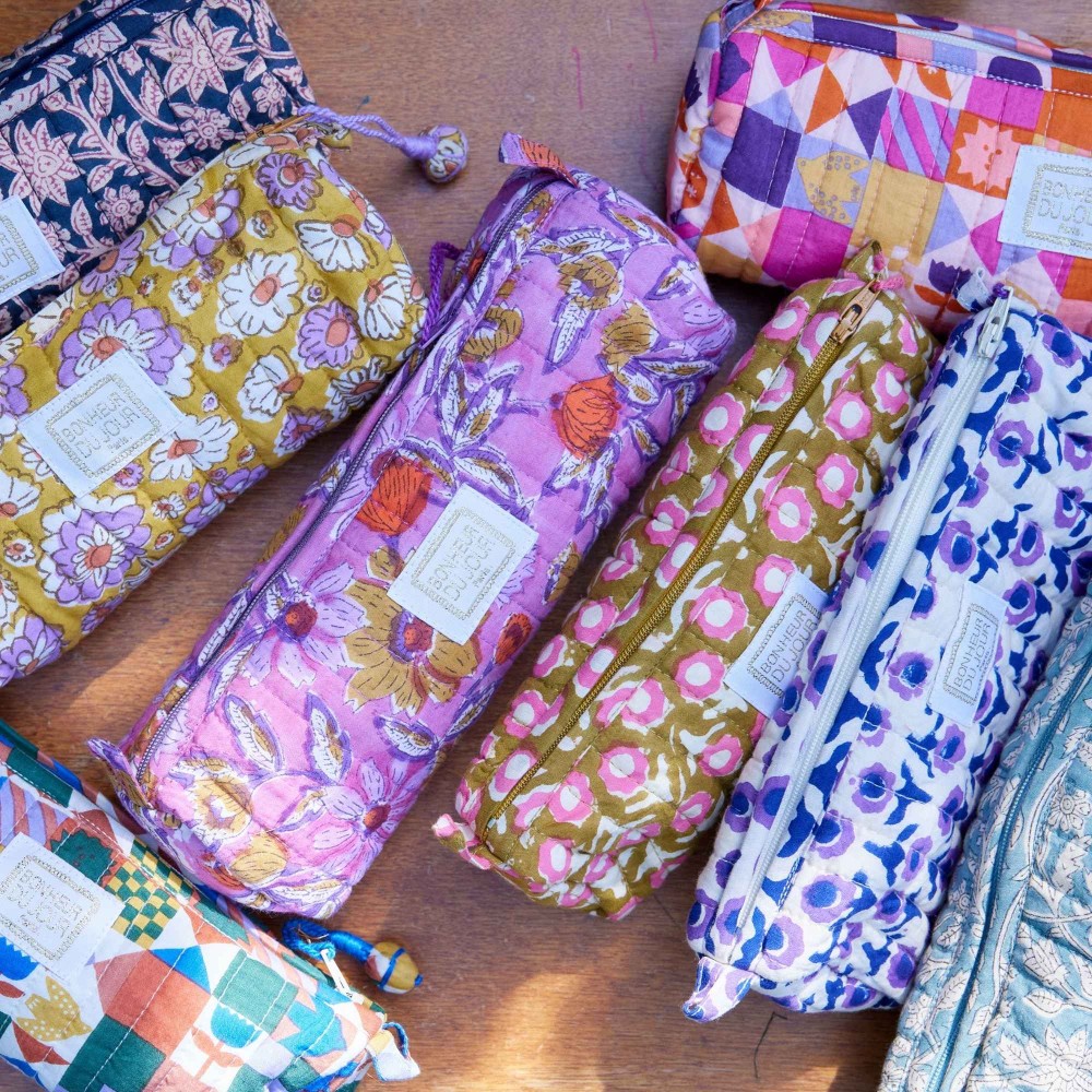 Several fabric zip pouches with colorful floral and geometric prints are arranged on a wooden surface. Each pouch has a small white sewn label.