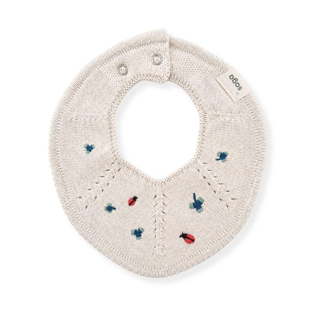 An off-white knitted baby bib with embroidered ladybugs and bees in red and blue, featuring a small brand tag that reads 'sodas'.