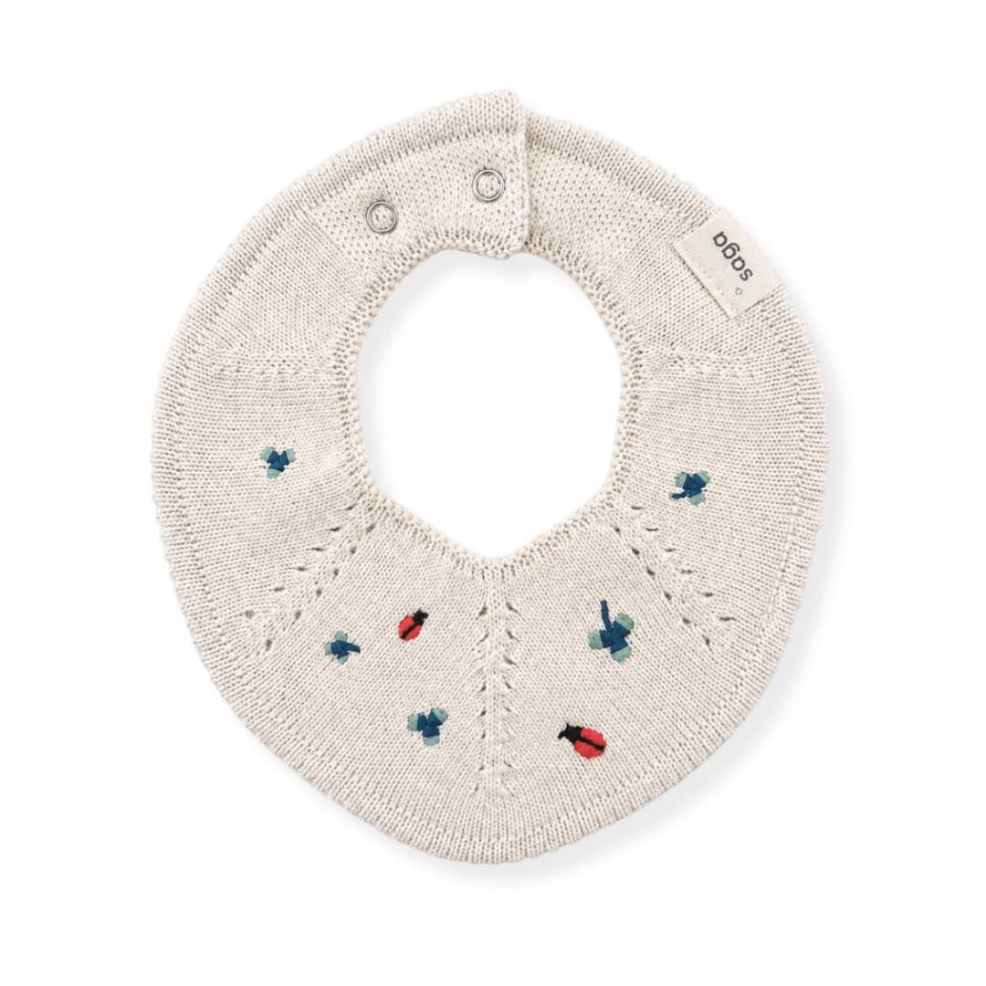 An off-white knitted baby bib with embroidered ladybugs and bees in red and blue, featuring a small brand tag that reads 'sodas'.