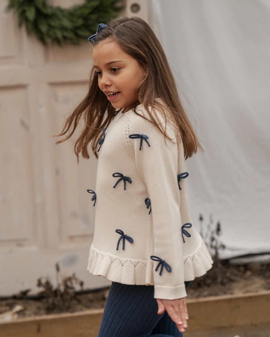 Young girl wearing a cream sweater with blue bow details outdoors.