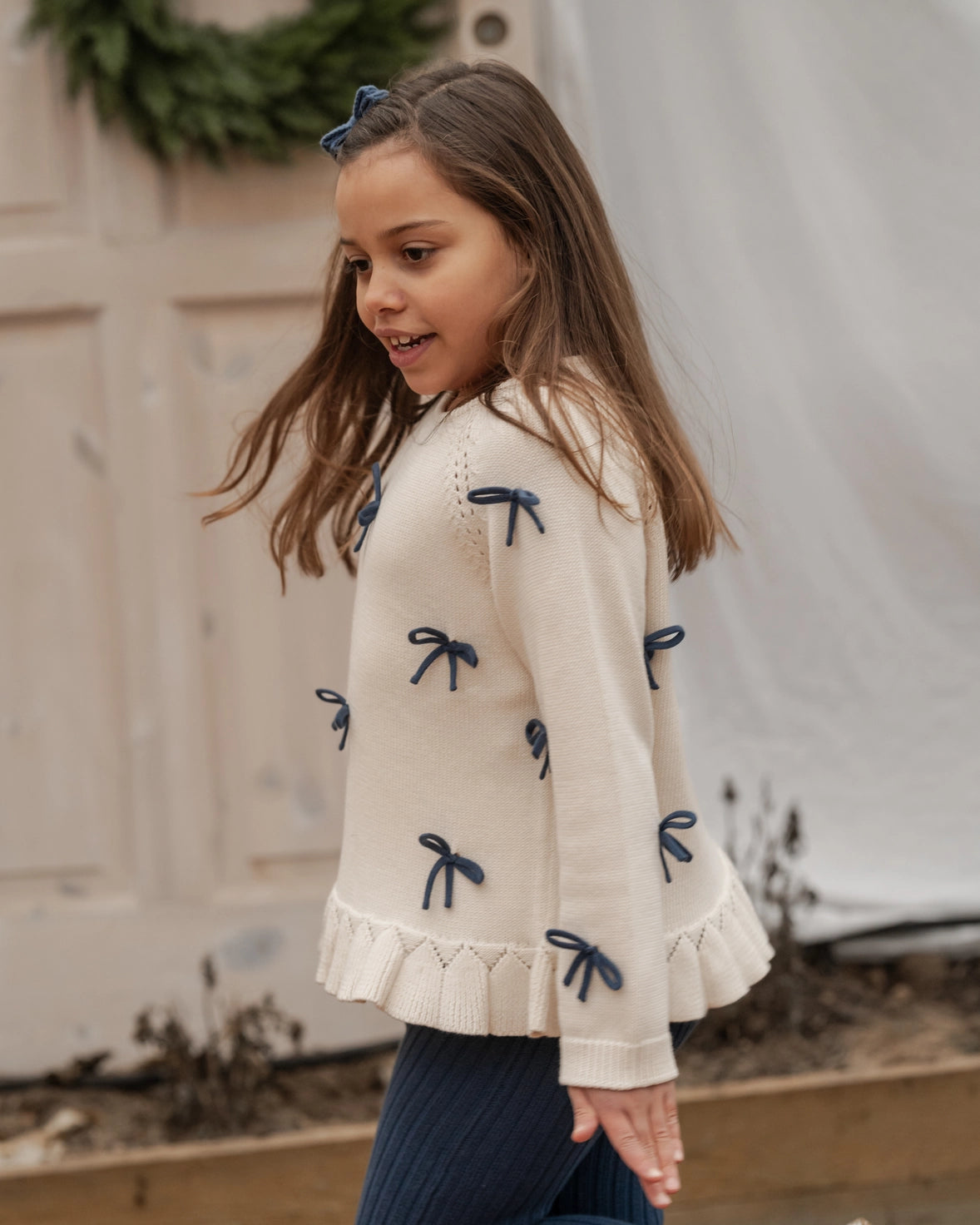 Young girl wearing a cream sweater with blue bow details outdoors.