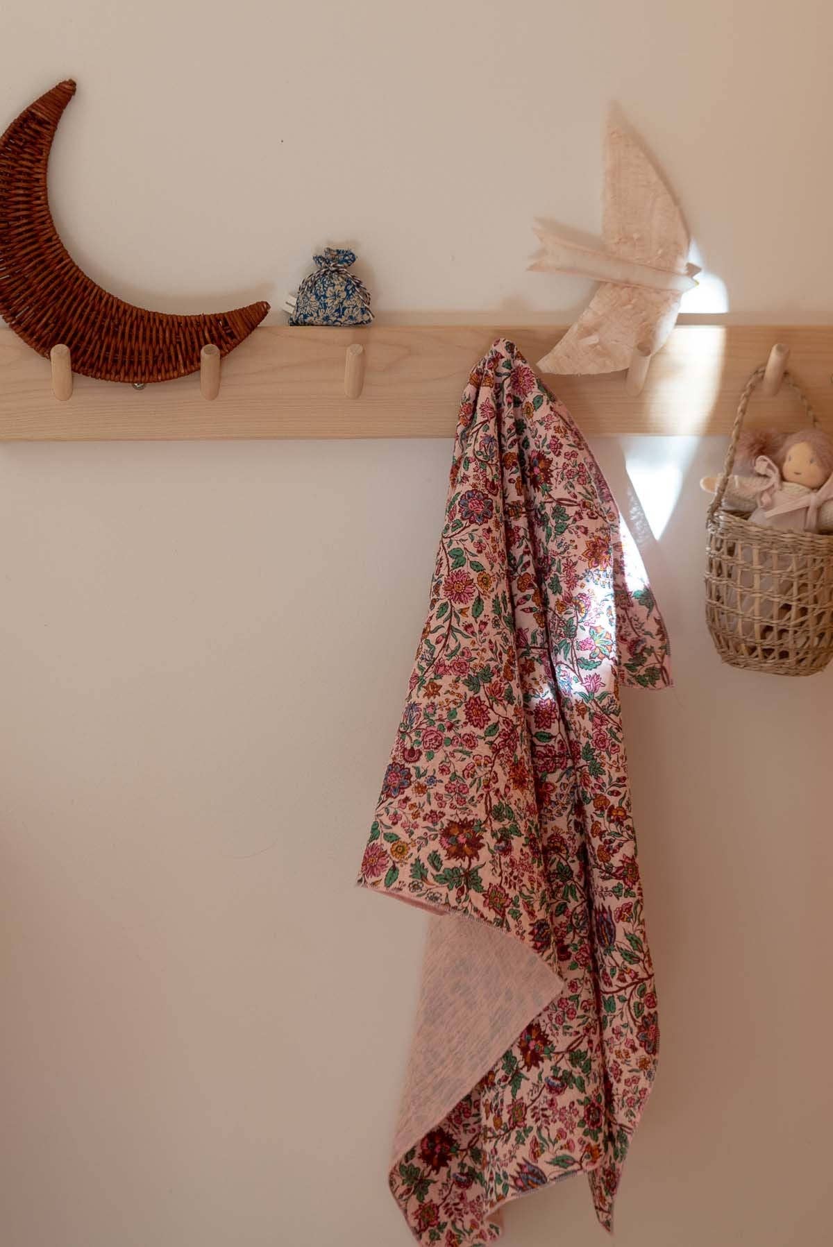 A floral patterned fabric throw blanket with a pink solid lining hangs from a light wood wall hook in a styled interior setting.
