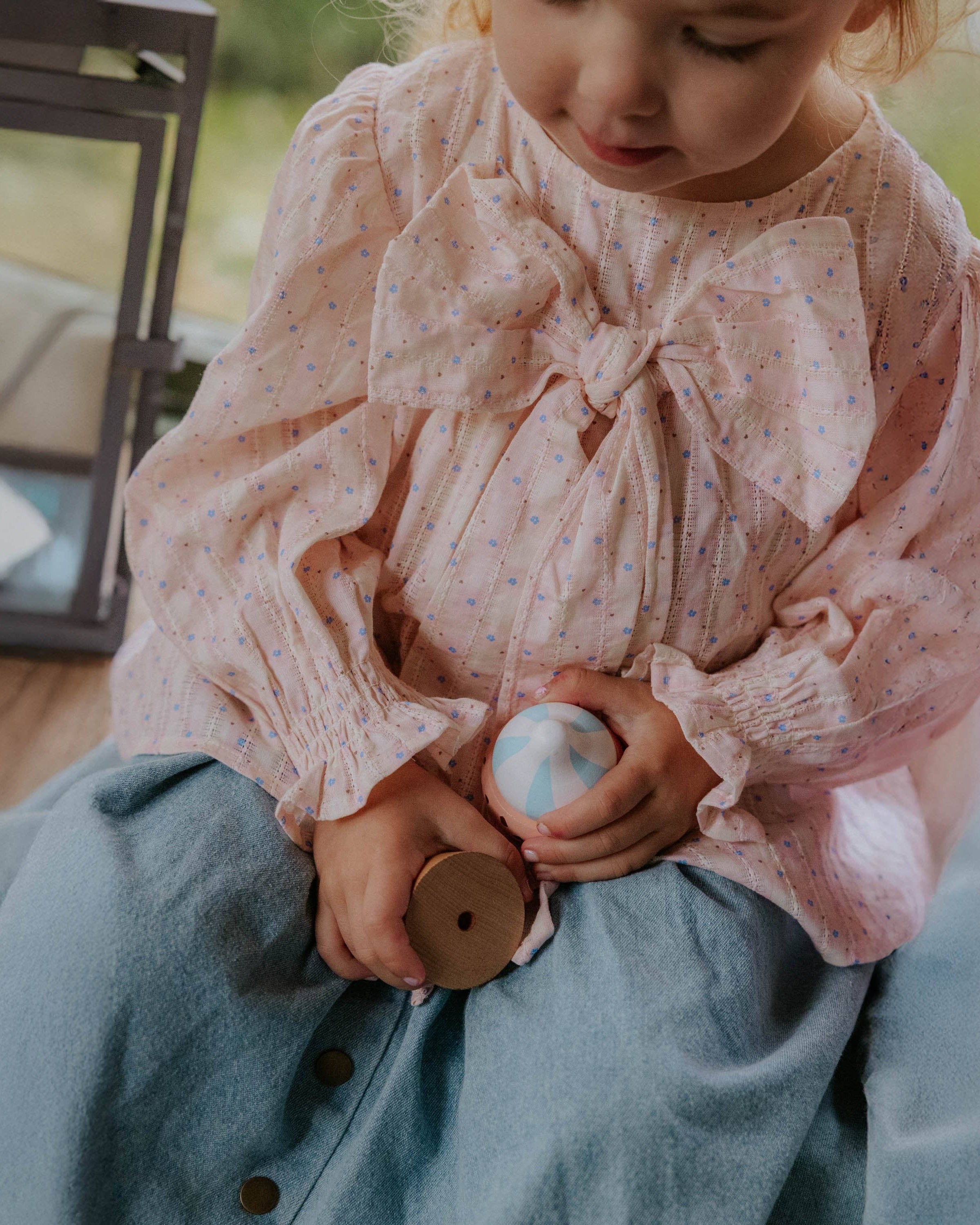 Child wearing a pink blouse with a large bow and blue pants, holding a wooden toy.