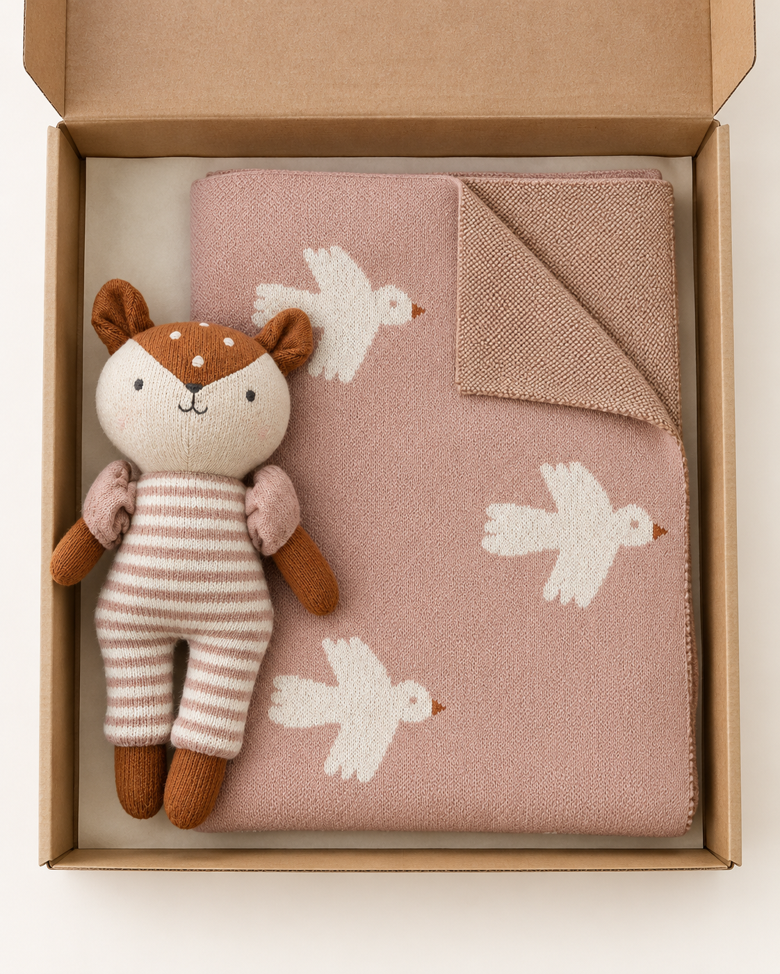 A baby gift set in a kraft paper box, featuring a dusty pink knit blanket with white bird patterns and a plush toy dressed as a deer in beige and brown striped clothing.