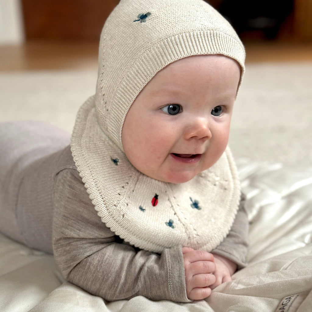 Baby wearing a beige knitted hat and scarf with embroidered designs on a light surface.