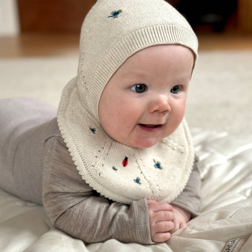 Baby wearing a beige knitted hat and scarf with embroidered designs on a light surface.