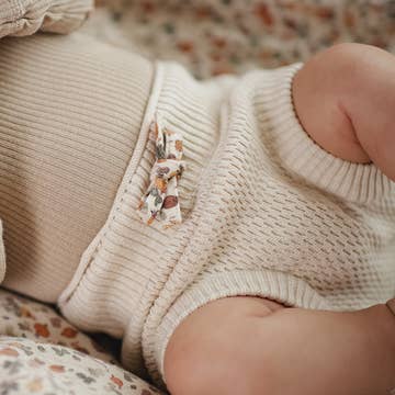 A close-up of a baby lying on a soft beige blanket, wearing a cream-colored waffle-knit romper with a small decorative animal applique at the waist.