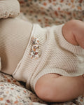 A close-up of a baby lying on a soft beige blanket, wearing a cream-colored waffle-knit romper with a small decorative animal applique at the waist.